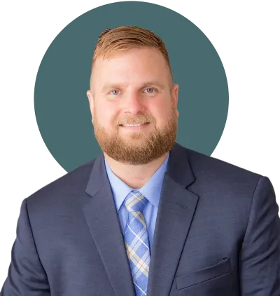 Professional headshot of a man with short blond hair and a beard, wearing a navy suit and blue plaid tie, smiling against a teal circular background.