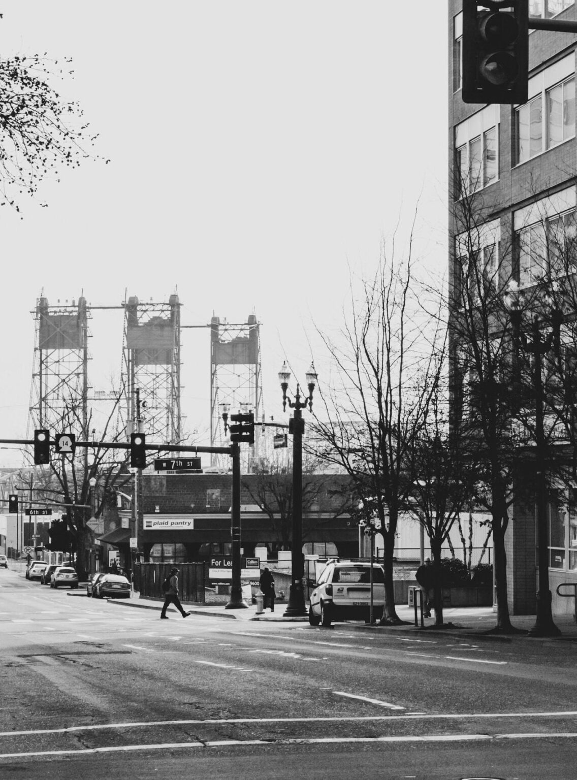 Black-and-white street scene with pedestrians crossing, cars parked, and a tall building on the right.